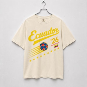May include: Cream-colored t-shirt with a soccer-themed design. The shirt features the word "Ecuador" in yellow script, a soccer ball graphic, the number '26', and the words "Soccer Club". A flag of Ecuador is also included.