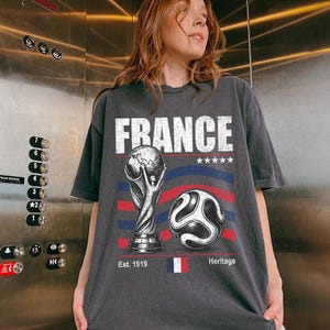 May include: A charcoal gray short-sleeve t-shirt featuring a graphic print with the text FRANCE, a world cup trophy, a soccer ball, five stars, and the French flag. The design includes the text Est. 1919 and Heritage with blue and red stripes.