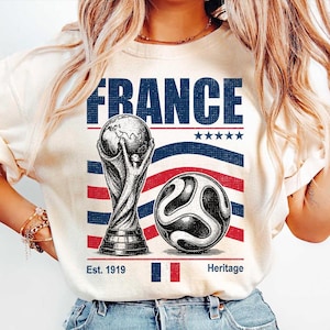 May include: A cream-colored t-shirt featuring a graphic with the word FRANCE in blue, a soccer trophy, a soccer ball, five stars, and the text Est. 1919 Heritage with a small French flag. The design uses blue and red stripes as a background element.