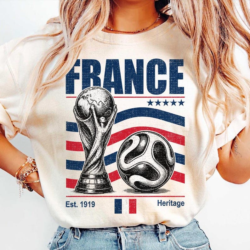 May include: A cream-colored t-shirt featuring a graphic with the word FRANCE in blue, a soccer trophy, a soccer ball, five stars, and the text Est. 1919 Heritage with a small French flag. The design uses blue and red stripes as a background element.
