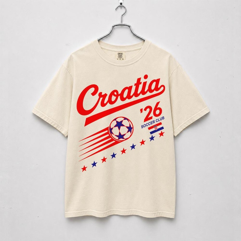 May include: A cream-colored short-sleeve t-shirt featuring a red graphic that reads Croatia '26 Soccer Club. The design includes a soccer ball with motion lines, the Croatian flag, and a diagonal row of alternating red and blue stars.