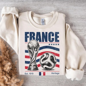 May include: A cream-colored crewneck sweatshirt featuring a graphic print with the word FRANCE in blue, a trophy illustration, a soccer ball, five blue stars, and red and blue wavy stripes. The text Est. 1919 and Heritage appear at the bottom.