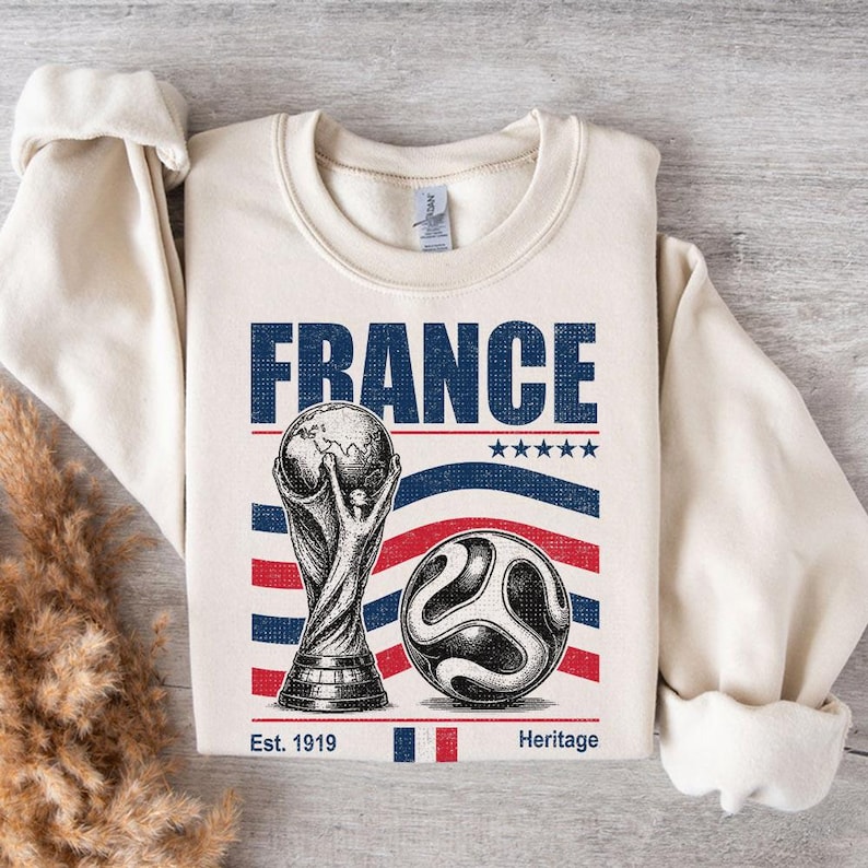 May include: A cream-colored crewneck sweatshirt featuring a graphic print with the word FRANCE in blue, a trophy illustration, a soccer ball, five blue stars, and red and blue wavy stripes. The text Est. 1919 and Heritage appear at the bottom.