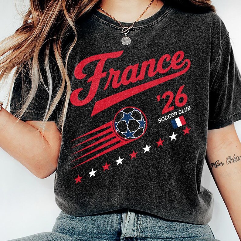 May include: Dark gray t-shirt with the word "France" in red script, the number "'26", and a soccer ball graphic with blue and white stars. The shirt also features the text "Soccer Club" and the French flag.
