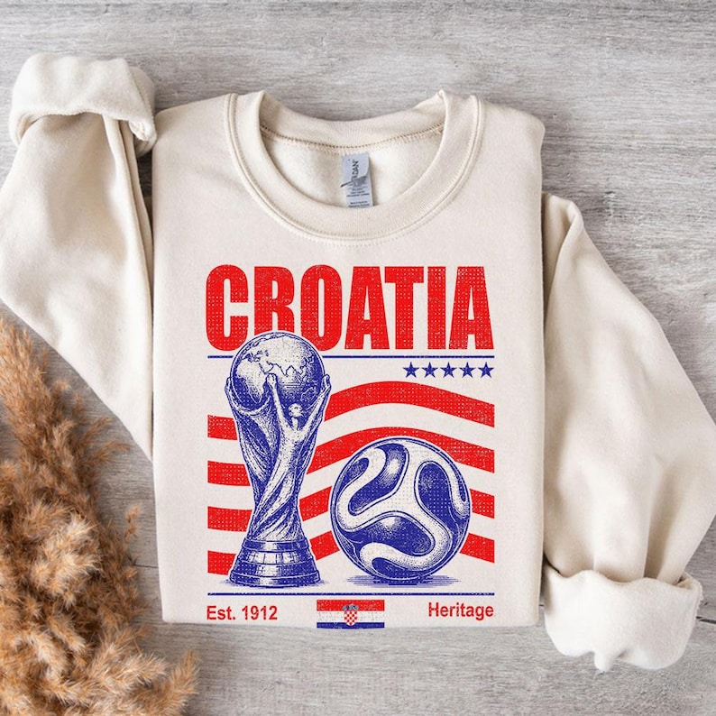 May include: A cream-colored crewneck sweatshirt featuring a graphic print with the word CROATIA in red letters. The design includes a blue illustration of a trophy, a soccer ball, five blue stars, red stripes, and a small Croatian flag with the text Est. 1912 Heritage.