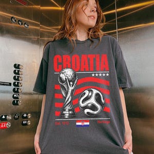 May include: A charcoal gray t-shirt featuring a red graphic print that reads CROATIA. The design includes a soccer trophy, a soccer ball, five white stars, and the Croatian flag with the text Est. 1912 Heritage printed in red below the flag.