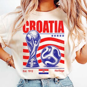 May include: A cream-colored t-shirt featuring a graphic with the word CROATIA in red, a blue trophy, a blue soccer ball, red stripes, five blue stars, the text Est. 1912, the text Heritage, and a small rectangular Croatian flag at the bottom center.