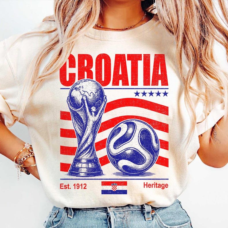 May include: A cream-colored t-shirt featuring a graphic with the word CROATIA in red, a blue trophy, a blue soccer ball, red stripes, five blue stars, the text Est. 1912, the text Heritage, and a small rectangular Croatian flag at the bottom center.