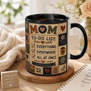Op de afbeelding: Een keramische mok met een zwarte handgreep en rand, voorzien van een patchwork-ontwerp met bloemen- en hartpatronen. De tekst luidt MOM TO-DO LIST: EVERYTHING, EVERYWHERE, ALL AT ONCE, met vinkjes en de hashtag #DOINGMYBEST op de voorzijde.