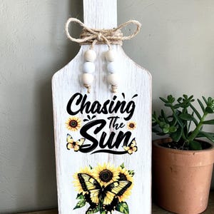 May include: White wooden cutting board with a handle, decorated with a sunflower and butterfly design. The text "Chasing the Sun" is printed on the board. The handle is adorned with a twine bow and wooden beads.