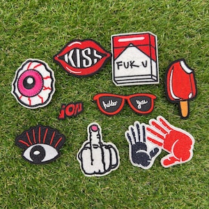 Può includere: Una collezione di toppe ricamate con labbra rosse e la scritta KISS, un pacchetto di sigarette rosso e bianco con la scritta FUK V, un bulbo oculare rosa, un ghiacciolo rosso, occhiali rossi con la scritta hello you, un occhio nero e due mani.