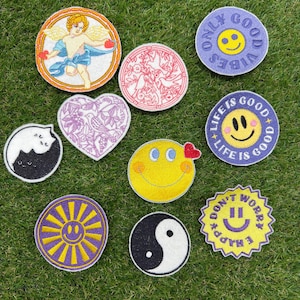 Puede incluir: Una colección de diez parches bordados con diversos diseños, incluyendo un querubín, un símbolo yin-yang, un gato, patrones florales y varias caras sonrientes con textos como Only Good Vibes, Life Is Good y Don't Worry Be Happy.