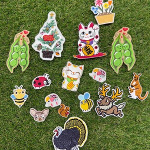 Puede incluir: Una colección de parches bordados con diversos diseños, incluyendo un árbol de Navidad, dos gatos de la suerte, un pavo, un canguro, un ciervo, abejas, una mariquita, una ardilla, un perro, una flor en maceta y dos vainas de guisantes.