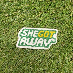 Puede incluir: Un parche bordado rectangular con fondo blanco y borde verde. El texto SHE GOT AWAY está cosido en una fuente negrita y estilizada. Las palabras SHE y AWAY son verdes, mientras que la palabra GOT es amarilla, todas con un contorno verde grueso.