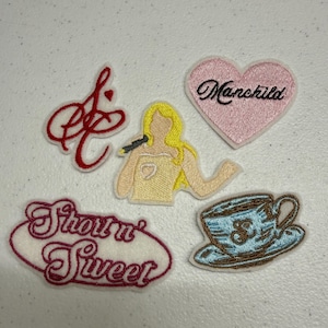 Puede incluir: Un conjunto de cinco parches bordados que incluye un monograma rojo en cursiva, una figura rubia sosteniendo un micrófono, un corazón rosa con el texto Manchild, una taza de té azul claro con plato y un parche ovalado blanco con el texto rojo Short n' Sweet.