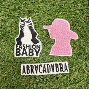 Puede incluir: Tres parches bordados: uno blanco con el texto negro WALK WALK FASHION BABY, uno rosa sólido con forma de persona con sombrero de ala ancha y uno rectangular blanco con el texto negro ABRYCADYBRA.