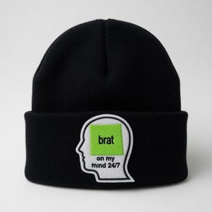 Pode incluir: Um gorro de malha preto com um emblema bordado branco em forma de perfil de cabeça humana. Dentro da forma da cabeça há um quadrado verde-limão brilhante com a palavra "brat" em texto preto, seguida pelo texto "on my mind 24/7" abaixo.