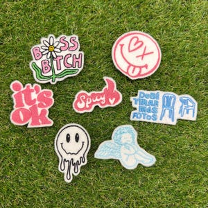 May include: A collection of seven embroidered patches featuring various designs: a daisy with the text BOSS BITCH, a pink smiley face, the text it's ok, the word Spicy, blue text reading Debí TIRAR más FOTOS with chair icons, a melting smiley face, and a blue angel.