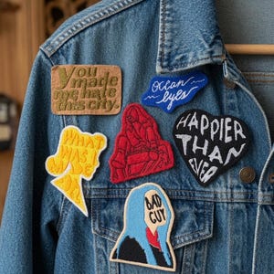 Puede incluir: Una chaqueta de mezclilla azul adornada con seis parches bordados. Los parches incluyen textos como "You made me hate this city", "ocean eyes", "WHAT WAS I MADE FOR", "HAPPIER THAN EVER" y "BAD GUY", junto a una ilustración de figura roja.