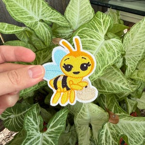 Puede incluir: Un parche bordado que muestra una abeja de dibujos animados con cuerpo amarillo, rayas negras y alas azul claro. La abeja tiene grandes ojos negros con pestañas largas y sostiene una pequeña cesta tejida de color beige en sus brazos.