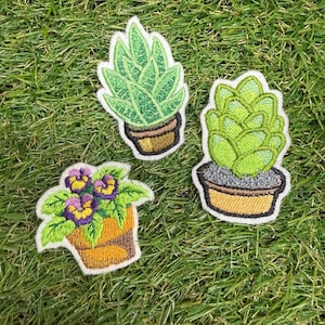 Puede incluir: Tres parches bordados con plantas en maceta. Un parche muestra una suculenta verde puntiaguda en una maceta marrón, otro una suculenta verde redondeada en una maceta marrón, y el tercero una planta con flores de pétalos morados y amarillos.