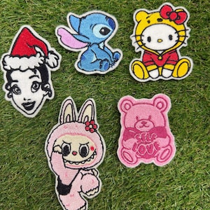 Puede incluir: Cinco parches bordados que muestran un personaje con gorro de Papá Noel, un alienígena azul, un gato con traje amarillo y rojo, un personaje rosa con orejas de conejo y un oso rosa que sostiene un corazón con el texto SELF LOVE bordado.