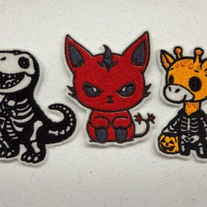 Puede incluir: Tres parches bordados con personajes de dibujos animados. A la izquierda, un esqueleto de dinosaurio en blanco y negro. En el centro, un diablo rojo con orejas y cola oscuras. A la derecha, una jirafa amarilla con un disfraz de esqueleto.