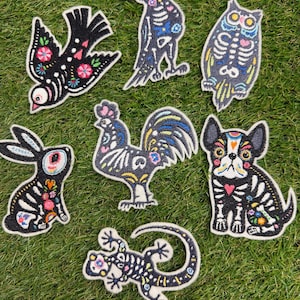 Puede incluir: Un conjunto de siete parches bordados con siluetas de animales en negro y detalles florales y esqueléticos coloridos. El set incluye un pájaro, un cuervo, un búho, un conejo, un gallo, un bulldog francés y una lagartija con costuras detalladas.