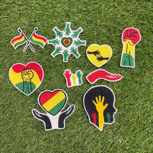 Puede incluir: Una colección de nueve parches bordados con diversos diseños, incluyendo banderas cruzadas, una estrella de manos verdes, un corazón amarillo con manos negras, un puño alzado, un corazón con un puño, manos pequeñas de colores y una silueta.