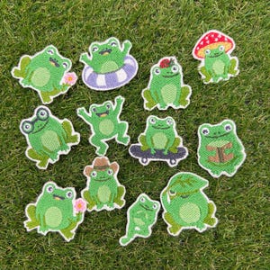 Puede incluir: Una colección de doce parches bordados con ranas verdes en varias posturas y accesorios, incluyendo una rana con una flor rosa, una rana en un flotador morado, una rana con sombrero de seta, una rana en monopatín y una rana leyendo.