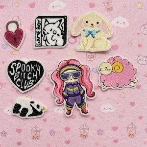 Puede incluir: Una colección de siete parches bordados que incluye un candado rojo en forma de corazón, una consola Game Boy en blanco y negro, un conejo amarillo con un lazo azul, un corazón negro con el texto Spooky Bitch Club, una oveja rosa, un panda y un personaje.