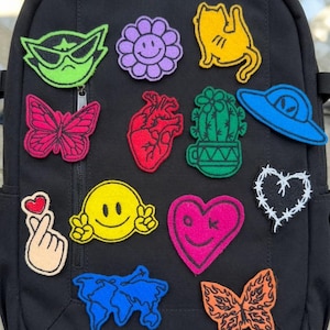 Puede incluir: Una mochila negra decorada con varios parches bordados, incluyendo una cara de gato verde, una flor morada, un gato amarillo, una mariposa rosa, un corazón anatómico rojo, un cactus verde, un OVNI azul, un signo de mano, una cara sonriente amarilla, un corazón rosa, un corazón de alambre de espino, un mapa mundial azul y una mariposa naranja.