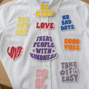 Puede incluir: Una camisa blanca de botones decorada con varios parches bordados de colores. Los parches incluyen textos como BE KIND, YES YOU CAN, LOVE, NO BAD DAYS, TREAT PEOPLE WITH KINDNESS, GOOD VIBES, LOVE, COOL y TAKE IT EASY con un símbolo de paz.