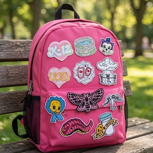 Puede incluir: Una mochila de color rosa brillante con varios parches bordados que incluyen un corazón con el texto K-12, un pozo de piedra, una cara de gato, un corazón con el texto void, una telaraña, un hongo, un pájaro azul, una polilla, un dragón y un gusano.