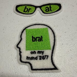 以下が含まれることがあります： 2つの刺繍ワッペン。1つはライムグリーンのレンズに黒い文字で「br」「at」と書かれたサングラスの形。もう1つは人間の頭部の白いシルエットで、ライムグリーンの四角形の中に「brat」「on my mind 24/7」という黒い文字が刺繍されています。