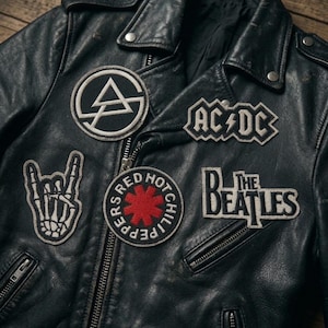 Puede incluir: Una chaqueta de cuero negra con varios parches bordados, incluyendo un logotipo circular de Linkin Park, un logotipo de AC/DC, una mano de esqueleto haciendo un signo de rock, un logotipo de Red Hot Chili Peppers y un parche con el texto The Beatles.