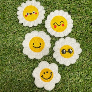 Puede incluir: Cinco parches bordados con forma de flor, con pétalos blancos y centros amarillos. Cada parche presenta una cara sonriente única con ojos y boca negros, algunos con mejillas rojas, realizados con hilo detallado sobre una base de tela blanca.