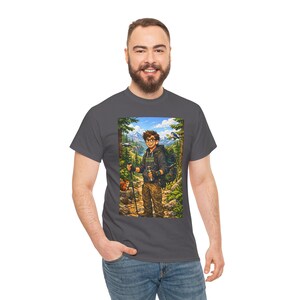 Puede incluir: Camiseta gris oscuro con una ilustración colorida de un excursionista en un entorno forestal. El excursionista lleva gafas, una mochila y un bastón de senderismo. La ilustración incluye una ardilla, montañas y el texto "Adventure Begins".