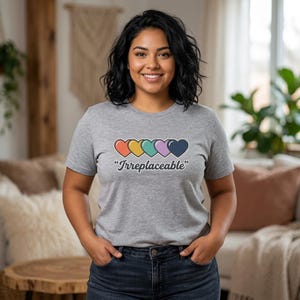 May include: A gray t-shirt with the word "Irreplaceable" and a row of five colorful hearts. The hearts are in shades of red, yellow, teal, purple, and navy blue. The shirt is made of a soft material.