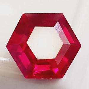 Könnte beinhalten: Ein facettierter, sechseckiger, roter Edelstein mit hohler Mitte. Der Edelstein hat scharfe, definierte Kanten und eine polierte Oberfläche, die das Licht reflektiert. Der Edelstein ist vor einem weißen Hintergrund platziert.