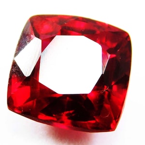 May include: A cushion-cut gemstone with a deep red colour. The gem has a large, open centre and multiple facets that reflect light. The gemstone is set against a white background.