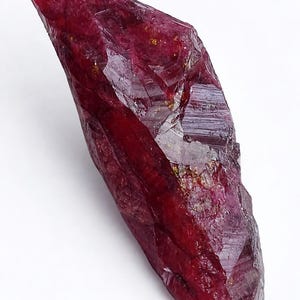 Peut inclure: Un cristal de rubis brut, non taillé, d'un rouge profond. La surface est rugueuse et texturée, avec des facettes visibles et une forme irrégulière. La pierre précieuse est présentée sur un fond blanc.