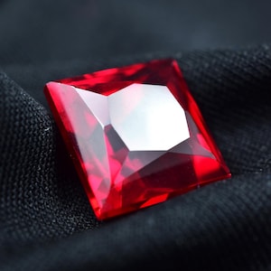 Peut inclure: Une pierre précieuse rouge vif, taillée en carré et à facettes. La gemme présente une surface centrale blanche et réfléchissante, sur un fond sombre et texturé. Les bords de la pierre sont nets et bien définis.