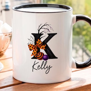 Puede incluir: Una taza de cerámica blanca con borde y asa negros, que presenta una letra K negra grande decorada con una telaraña, un lazo naranja con puntos negros y pequeñas esferas. El nombre Kelly está impreso en letra cursiva negra debajo.