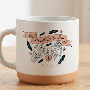 May include: A white ceramic mug with a speckled finish and a terracotta-colored base. The front features a graphic of two skeleton hands holding a heart-shaped lock, a banner with the text 'Til Death Do Us Part', and the date '06.25.26'.