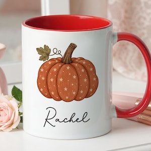 May include: A white ceramic mug with a bright red interior and handle. The exterior features a graphic of an orange pumpkin with a small green leaf and a pattern of tiny stars. The name Rachel is printed in black cursive script below the pumpkin.