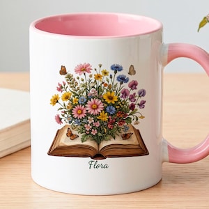 Peut inclure: Un mug en céramique blanche avec un intérieur et une anse rose pâle. L'extérieur présente une illustration colorée d'un bouquet de fleurs sauvages sortant d'un livre ouvert, avec de petits papillons et le mot Flora écrit en noir.
