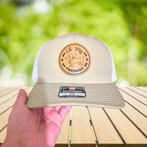 May include: Tan and white trucker hat with a circular patch featuring a dachshund illustration and the text "Oh My Gweenies!". The hat has a curved brim and an adjustable strap. The Richardson brand tag is visible.