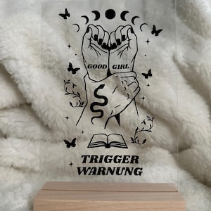Könnte beinhalten: Klarer Acrylschild mit Holzsockel. Das Schild zeigt eine schwarze Grafik von zwei Händen, einer Schlange, Schmetterlingen und den Worten "GOOD GIRL" und "TRIGGER WARNUNG". Das Schild ist zum Aufstellen konzipiert.