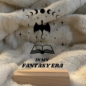 Könnte beinhalten: Klarer Acrylschild mit einem schwarzen Drachen, Mondphasen, Sternen und einem offenen Buchdesign. Der Text "IN MY FANTASY ERA" befindet sich unten. Das Schild steht auf einem Holzsockel.